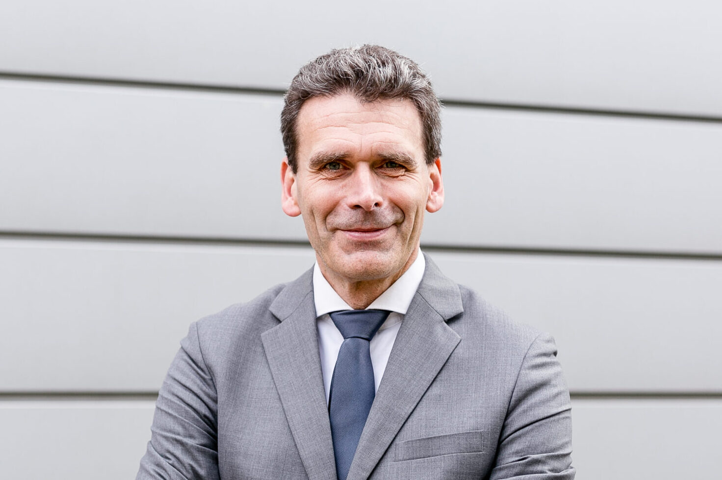 Prof. Dr. Frank Dellmann. Foto: FH Münster Das Bild zeigt Herrn Prof. Dr. Frank Dellmann im Porträt. Er lächelt, trägt einen grauen Blazer und die Arme verschränkt. Der Bildhintergrund ist etwas heller grau.