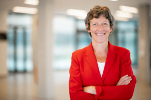 Das Bild zeigt Frau Prof. Dr. Susanne Staude im Porträt. Sie lächelt und trägt einen roten Blazer. Der Bildhintergrund ist unscharf.