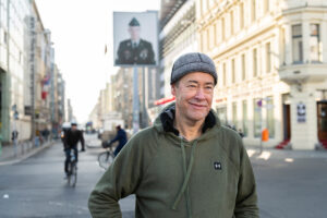 Thomas Krüger, Präsident der Bundeszentrale für politische Bildung, steht vor dem ehemaligen Grenzübergang Checkpoint Charlie in Berlin. Er trägt ein olivgrünes Sweatshirt und eine graue Mütze. Im Hintergrund sind auf der Straße zwei Radfahrer und an einem Masten ein Bild eines Soldaten unscharf zu erkennen.