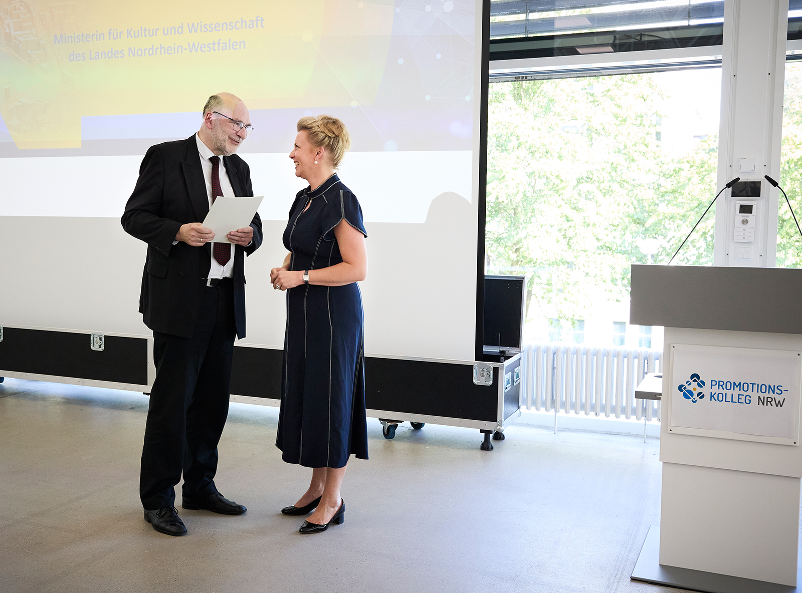 Im Bild sieht man NRW-Wissenschaftsministerin Ina Brandes (rechts im Bild) und Prof. Dr. Martin Sternberg nebeneinander stehen. Sie sind im Gespräch. Martin Sternberg hält eine Urkunde in der Hand. Rechts steht ein Pult auf dem ein Schild mit der Aufschrift "Promotionskolleg NRW" angebracht ist. Foto: Heike Fischer, Köln