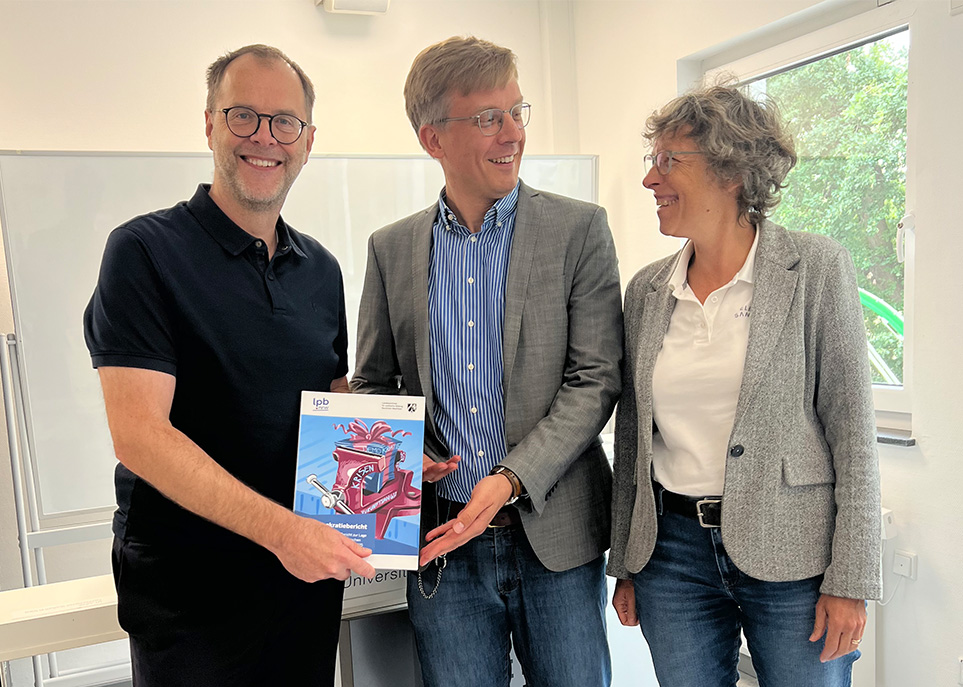 Im Bild von links nach rechts stehen Prof. Dr. Bernd Kriegesmann (Vorsitzender der Hochschulen NRW), Dr. Arne Hordt (Referent für Grundsatzangelegenheiten der Landeszentrale für politische Bildung Nordrhein-Westfalen) und Prof. Dr. Susanne Staude (stellvertretende Vorsitzende der Hochschulen NRW). Während sich Staude und Hordt anschauen, präsentiert Kriegesmann den Demokratiebericht in Richtung Kamera.