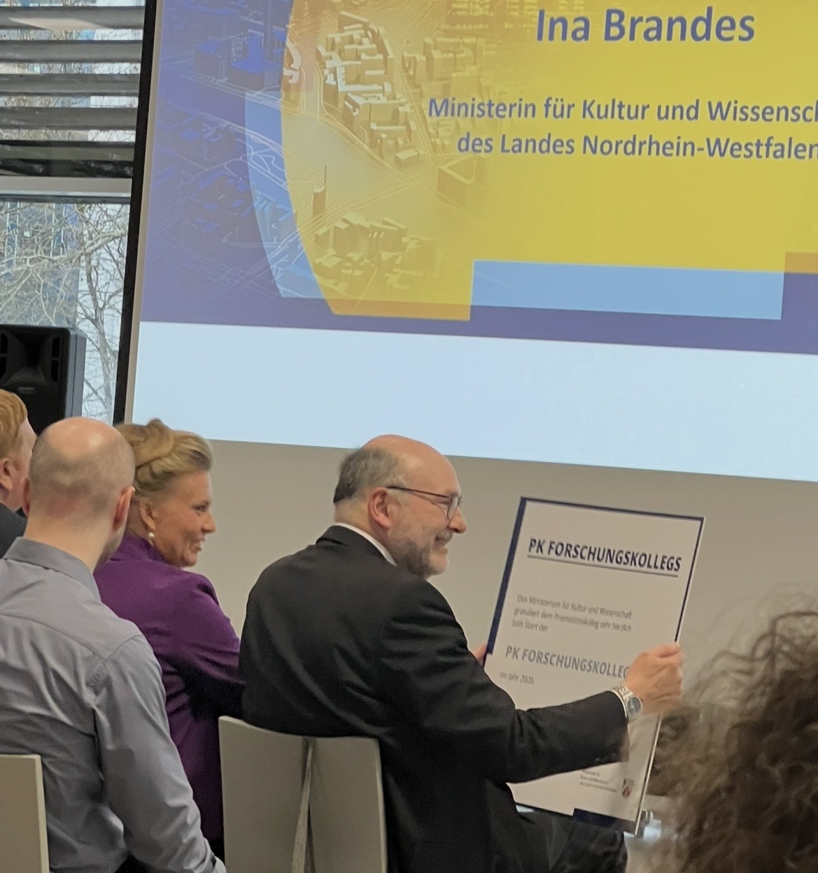 Auf dem Foto sieht man Menschen während einer Veranstaltung auf Stühlen sitzen. Halb von hinten ist Prof. Dr. Martin Sternberg zu erkennen, der eine große symbolische Förderzusage in Händen hält. Neben ihm sitzt NRW-Wissenschaftsministerin Ina Brandes. Beide lächeln.